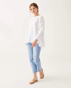 A woman with wavy brown hair models the MERSEA - CATALINA SLUB TEE—an oversized, long-sleeved white top with thin gray stripes—by MERSEA. She pairs it with blue jeans and stands against a plain, light background, hands behind her back.