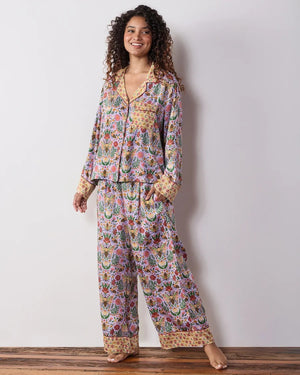 A person wearing the PRINTFRESH - BEE TAPESTRY ECO SATIN SOHO SET IN LAVENDER, featuring vintage-inspired bee and floral vine prints, holding a flower behind their back in a garden.