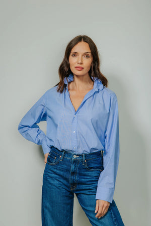 A woman with wavy brown hair wears the KMJ - Quarantina Shirt in Chambray, its feminine ruffle collar and relaxed fit highlighted as she stands with one hand in her pocket against a plain light gray background, looking confidently at the camera.