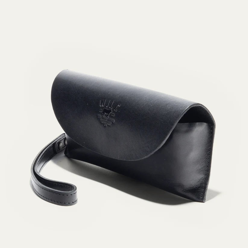 The WILL LEATHER GOODS - EYEGLASS CASE IN BLACK is a handcrafted Italian leather case with a flap closure, embossed logo on the front, and an attached wrist strap on the left, shown against a plain light background.