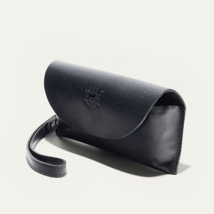 The WILL LEATHER GOODS - EYEGLASS CASE IN BLACK is a handcrafted Italian leather case with a flap closure, embossed logo on the front, and an attached wrist strap on the left, shown against a plain light background.