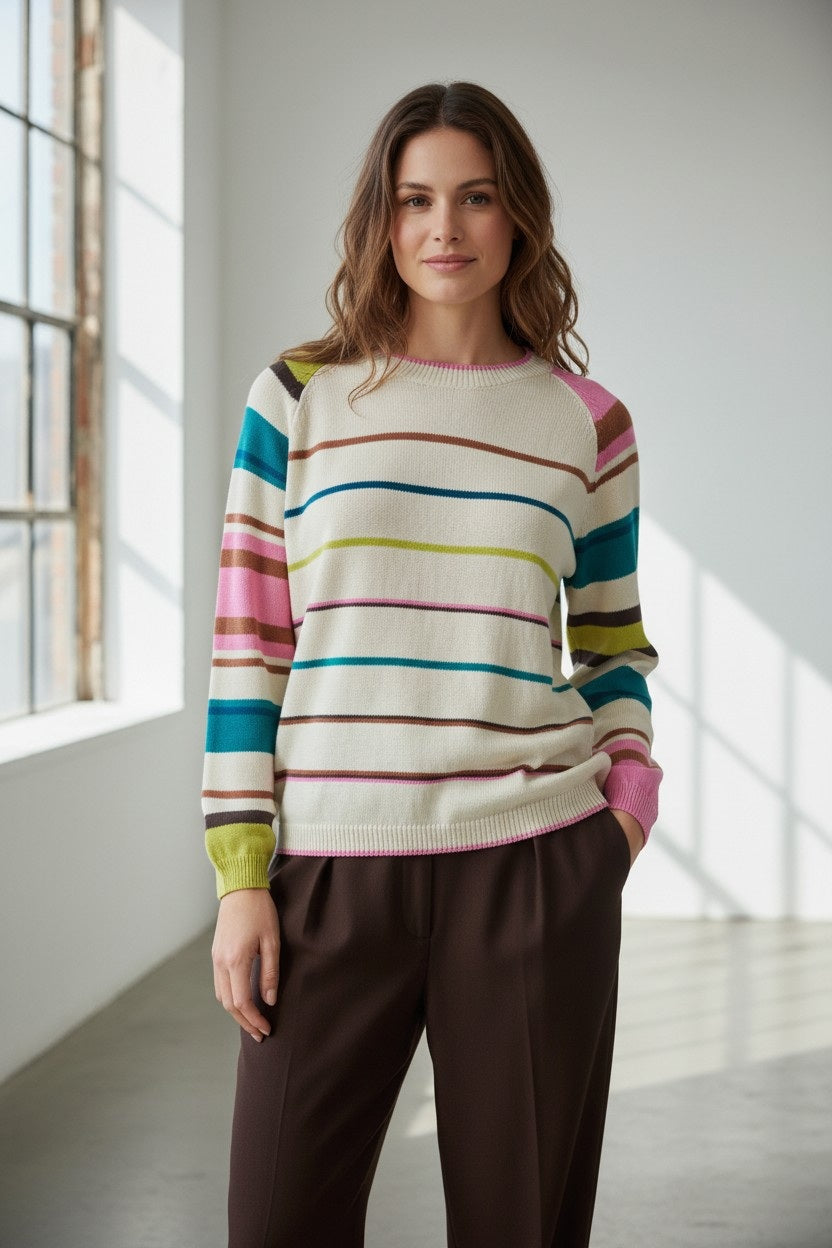 A woman stands indoors, smiling softly as sunlight streams through large windows and casts patterns on the floor. She wears the FLOOR - STRIPED COTTON SWEATER by FLOOR, its cream base adorned with colorful horizontal stripes, paired with brown pants.