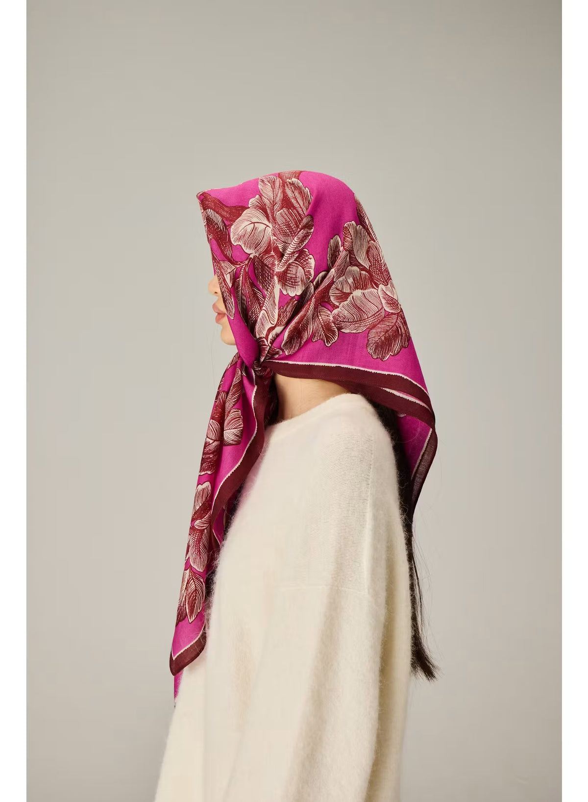 A woman with straight black hair wears a cream sweater and the INOUI EDITIONS - ARBORI Square Wool Scarf in Fuschia by INOUI, crafted from 100% wool and featuring a leaf pattern, draped around her neck against a plain light background.