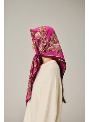 A woman with straight black hair wears a cream sweater and the INOUI EDITIONS - ARBORI Square Wool Scarf in Fuschia by INOUI, crafted from 100% wool and featuring a leaf pattern, draped around her neck against a plain light background.