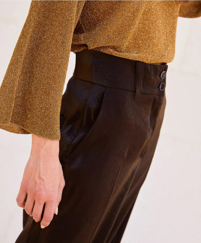 A person stands against a light-colored stone wall, wearing a loose-fitting, long-sleeved brown top with bio-galalite buttons paired with the high-waisted, wide-leg MANHATTAN PANT from MOMONI. They have long hair and are looking directly at the camera. The background is simple and uncluttered.