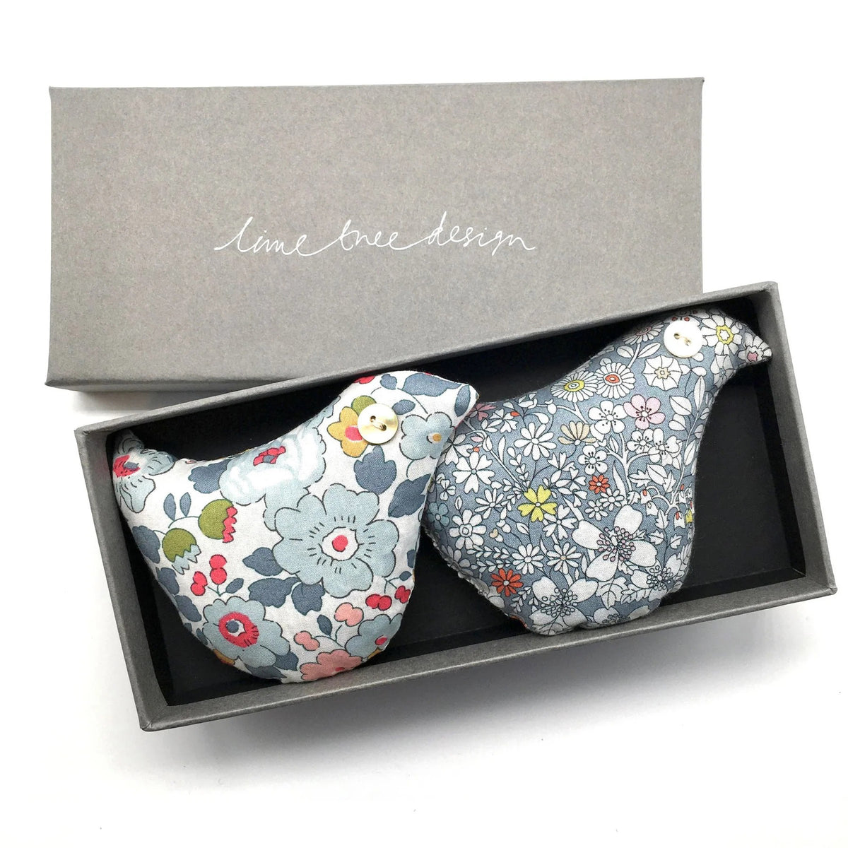 A grey gift box with the &quot;Lime Tree Collection Ltd&quot; logo on the lid contains two BIRDS - SONG BIRD SACHET made from Liberty Tana Lawn fabric. Each sachet showcases unique floral patterns: one featuring larger blue and pink flowers, and the other decorated with smaller multicolored flowers, evoking the fragrant lavender of Provence.
