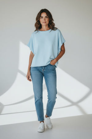 A woman with wavy brown hair stands in a bright room, smiling softly and wearing the FRANK & EILEEN Olive Original Capelet in Dive, paired with blue jeans and white sneakers. One hand is in her pocket.