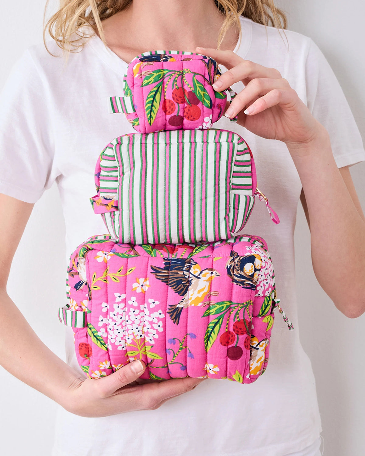 A person in a white shirt holds the PRINTFRESH - BIRD &amp; BLOSSOM THREE POUCH SET by PRINTFRESH, featuring three quilted makeup pouches with colorful floral and striped patterns, nested from smallest to largest.