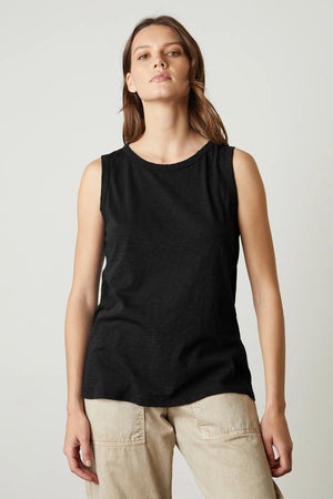 A person with shoulder-length brown hair is wearing the VELVET BY GRAHAM & SPENCER - TAURUS TANK IN COTTON SLUB and light-colored pants. They are standing in front of a plain white background, looking slightly upward with a neutral expression.