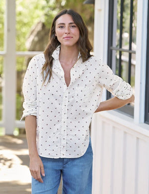 A woman with long wavy hair stands sideways on a porch, looking down, wearing the FRANK & EILEEN EILEEN Relaxed Button Up in Italian Cotton Palm Trees and blue pants. One hand rests in her pocket as the shirt’s relaxed fit drapes effortlessly, enhanced by bust-flattering button placement for subtle shape. The background is bright and green.