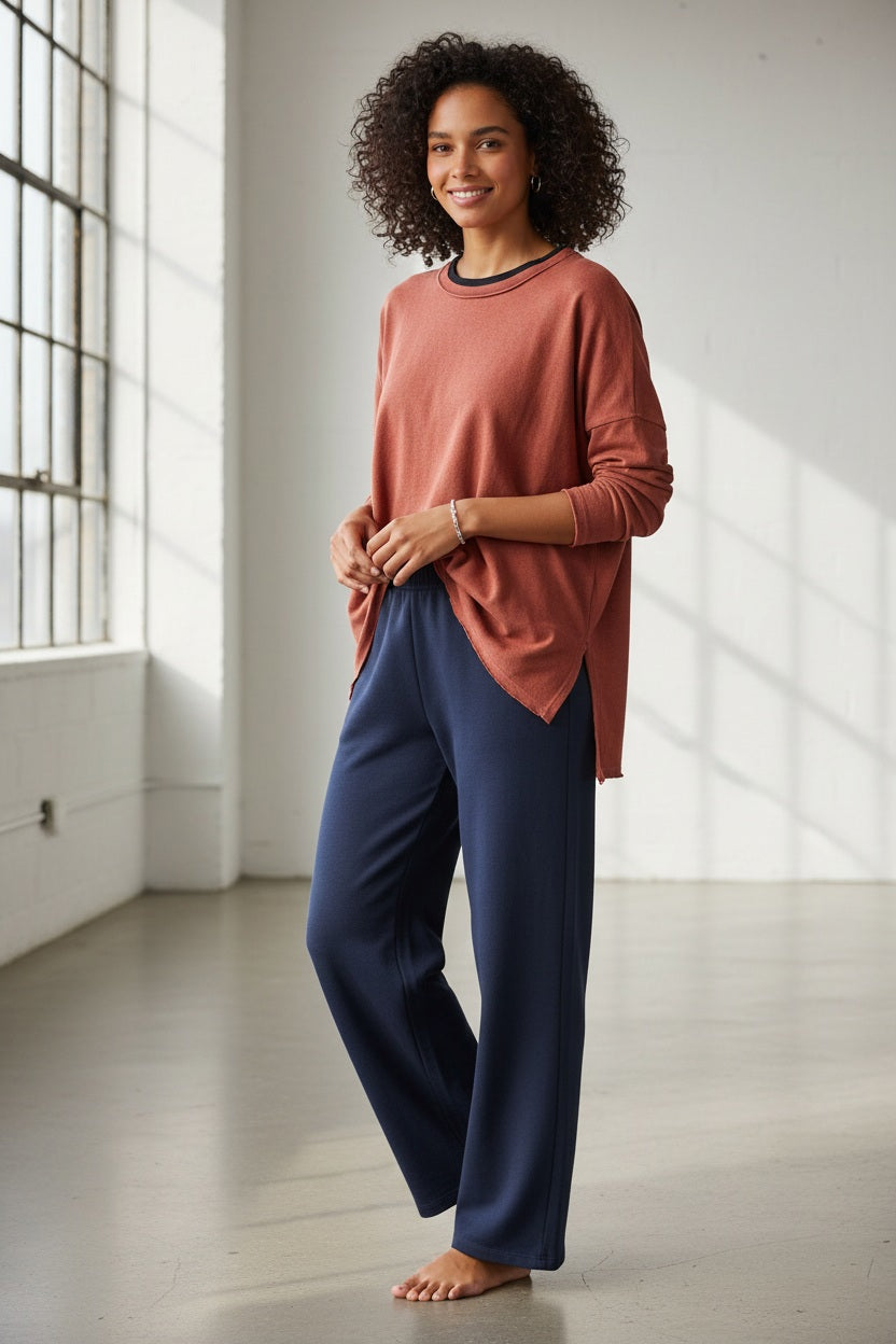 A woman with long brown hair wears the MERSEA Catalina Rustic Tee in Cayenne, paired with dark blue pants. She stands against a plain light background, resting her chin on her hand and gazing at the camera.