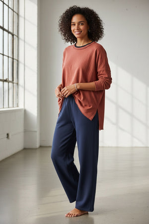 A woman with long brown hair wears the MERSEA Catalina Rustic Tee in Cayenne, paired with dark blue pants. She stands against a plain light background, resting her chin on her hand and gazing at the camera.