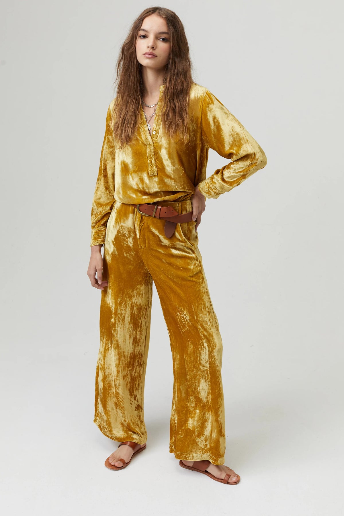 A person with long wavy hair stands against a plain background wearing a mustard yellow, pure silk velvet long-sleeve top and matching, high-waisted LANHTROPY - THE LUNA VELVET PANT by LANHTROPY. They are accessorized with layered necklaces, a brown belt, and flat sandals.