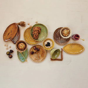 Four CREATIVE COOP DEBOSSED STONEWARE SCALLOPED PLATES with botanical designs and scalloped edges in brown, orange, green, and yellow, arranged in a square on a white background.