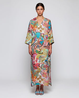 A woman in a MIRTO 1956 Multi Color Paisley Kaftan Style Dress with wide sleeves and V neckline, her hair in a low bun, wears light blue embellished-heel shoes against a plain white background.