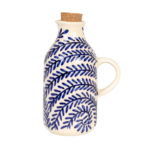 The HAND-PAINTED STONEWARE OIL CRUET WITH CORK STOPPER by CREATIVE COOP features blue leaf patterns and a handle, making it a perfect addition to rustic kitchen decor. Shown on a white background with the cork beside it.