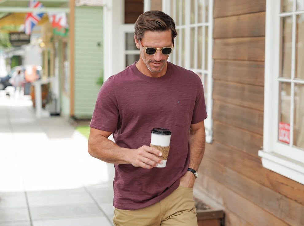 A person wearing the CRIQUET Slub Cotton Short Sleeve Pocket T-Shirt in Jam Band, featuring a small chest pocket and a subtle blue logo. The blurred greenery and wood background perfectly complement this vintage-inspired CRIQUET shirt, creating an effortlessly classic look.