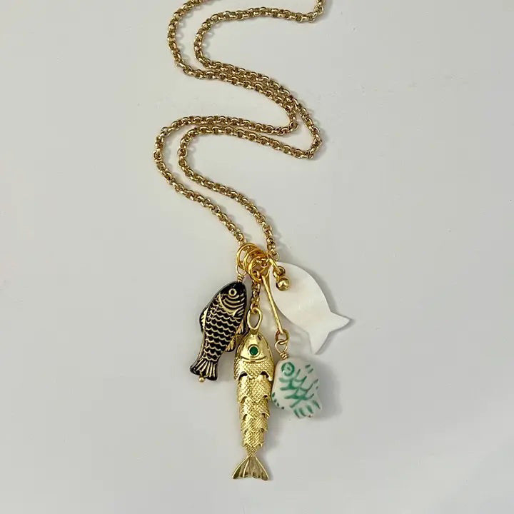 The SANDRINE FRANCE STUDIO - CORDELIA NECKLACE features handmade gold chain with four pendants—a black fish, a gold fish, white sea glass-like piece, and a ceramic School of Fish—beautifully displayed on a plain white surface.