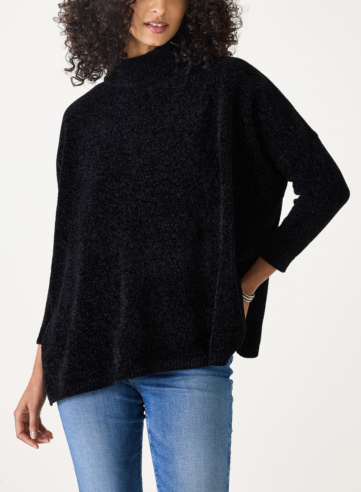 Wearing the MERSEA - CATALINA EVERSOFT CHENILLE SWEATER in black with light blue jeans, a curly-haired person stands against a plain light background, one hand on their hip.
