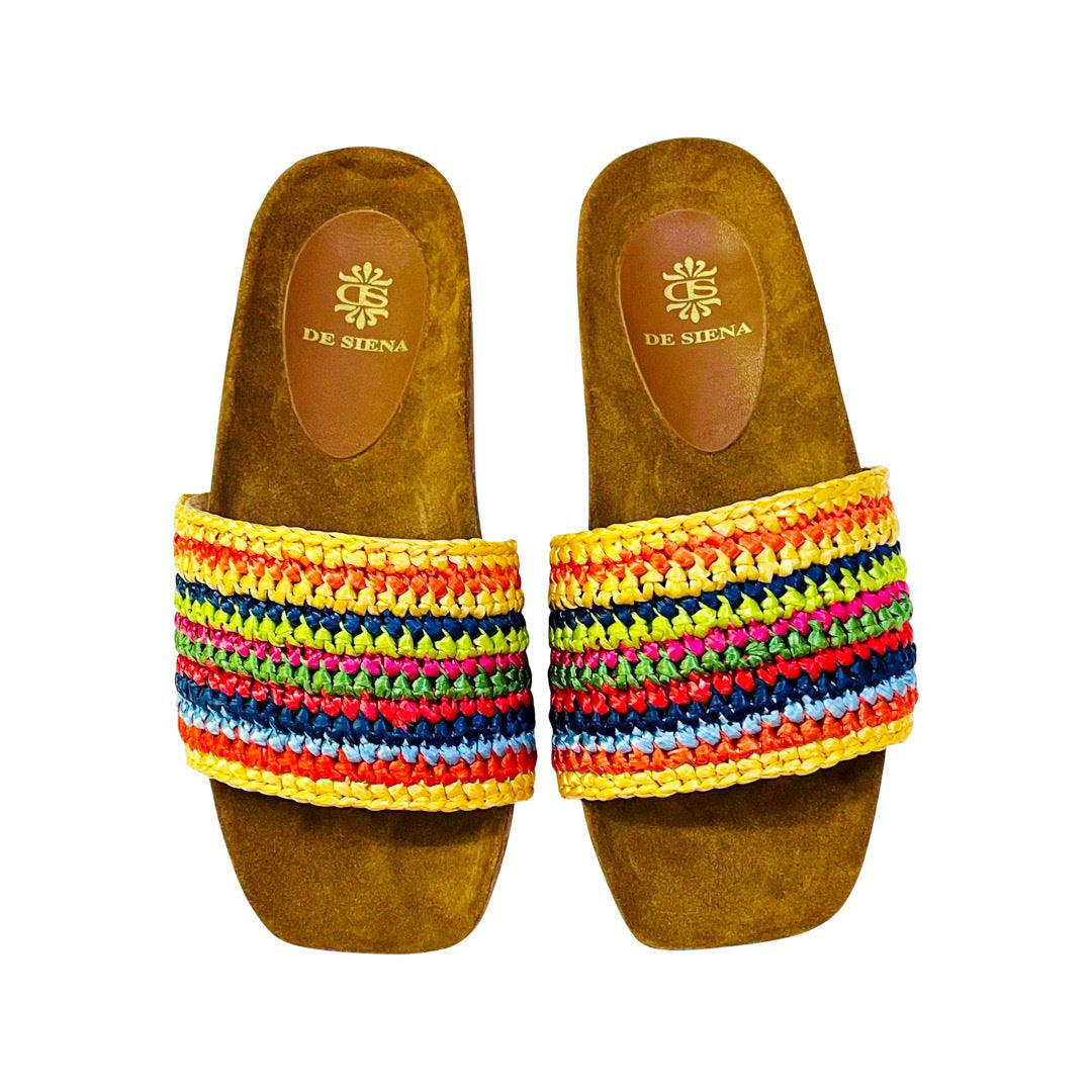 A pair of DA SIENA - LUCILLA RAFIA SLIDE sandals by DE SIENA features tan-colored soles with a raffia knotted top, adorned with multicolored stripes in shades of green, blue, brown, and yellow. The eco-style footbed is tan and showcases the brand's name and logo printed in gold, making it perfect for your summer style.