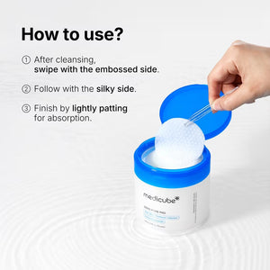 A hand holds a round, textured exfoliating pad above an open jar of KOSMIC KBEAUTY SKINCARE MEDICUBE - ZERO PORE PAD 2.0, 100ML, with its blue lid set aside on a plain white background.