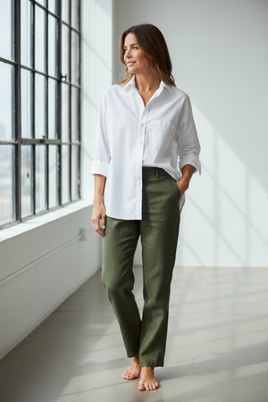 A woman smiles while leaning against a beige pillar, wearing the FRANK & EILEEN Oona Continuous Sleeve Button Up Shirt in Regenerative Cotton White and blue jeans, with one hand in her pocket. Neutral-toned artwork and furniture are visible in the background.
