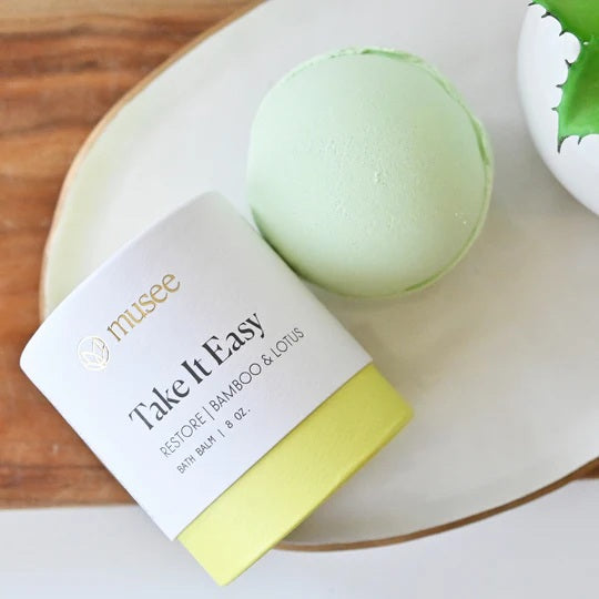 A pale green Musee - Take It Easy Bath Balm rests beside its cylindrical container, which is topped in white with a yellow base and labeled “MUSEE TAKE IT EASY BATH BALM 8 OZ.” The packaging highlights handcrafted bath products from Musee, created to deliver a naturally soothing bathing experience.