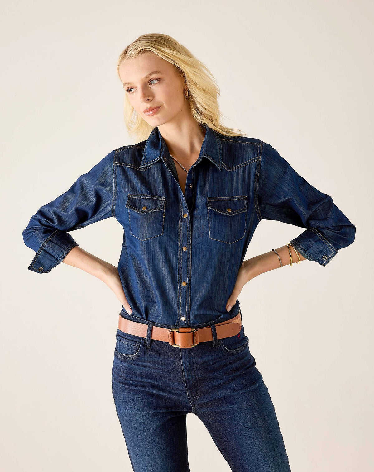 A woman stands indoors by a window, smiling and wearing the MERSEA MARGOT BUTTON UP SHIRT from MERSEA, featuring pearl snaps and a tailored fit with rolled-up sleeves. She pairs the dark denim shirt with matching jeans and a brown belt, while her dark hair falls in loose waves.
