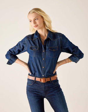 A woman stands indoors by a window, smiling and wearing the MERSEA MARGOT BUTTON UP SHIRT from MERSEA, featuring pearl snaps and a tailored fit with rolled-up sleeves. She pairs the dark denim shirt with matching jeans and a brown belt, while her dark hair falls in loose waves.