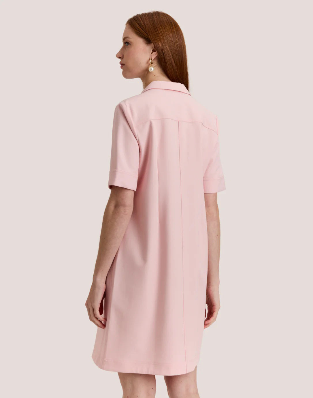 A woman with long, straight auburn hair wears the HINSON WU Bridget Short Sleeve Traveler's Twill Dress in blush pink, featuring a collar and gold front zipper, standing against a plain light background.