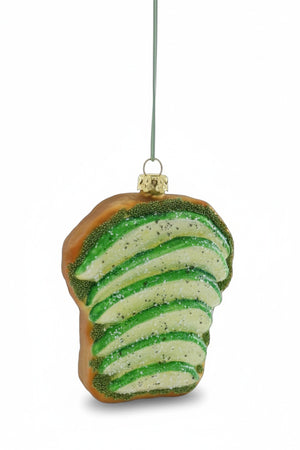 The AVOCADO TOAST ORNAMENT by CODY FOSTER is a decorative glass piece shaped like toast topped with avocado slices, designed with glitter and suspended from a string. It features a glossy finish that artfully combines green and brown hues, while its transparency enhances its elegant appeal.