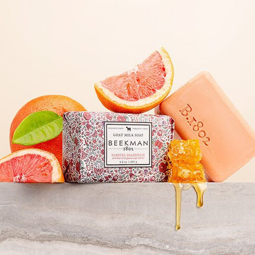 The BEEKMAN - STA ELEMENTS HONEYED GRAPEFRUIT 3.5 SOAP is a rectangular exfoliating goat milk bar wrapped in white paper with a gray checkered pattern, featuring orange grapefruit slices and honeycomb illustrations for a natural glow.