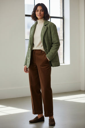 A woman with short, wavy dark hair stands indoors in a bright room with large windows, wearing the FRANK & EILEEN Quilted Belfast Jacket in Vintage Green over a white turtleneck, brown pants, and dark loafers. The light green jacket is made from Italian peached twill.