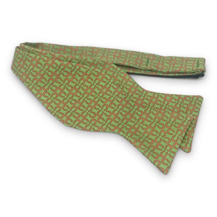 A handmade silk bow tie by Bird Dog Bay, the Peter-Blair Ringers design in green and red with a repeating circular pattern, features adjustable neck sizing and is displayed on a white background.