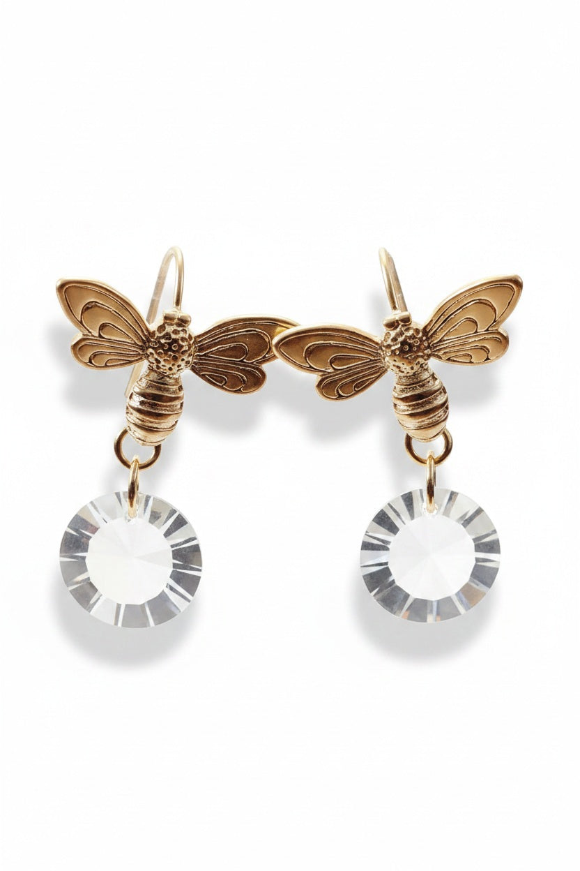 The VERDIER - HONEY BEE FROP EARRINGS WITH SWAROVSKI MOON DISC EARRINGS by VERDIER, featuring gold bee shapes with clear Swarovski crystal drops, are displayed hanging on the rim of a clear glass.