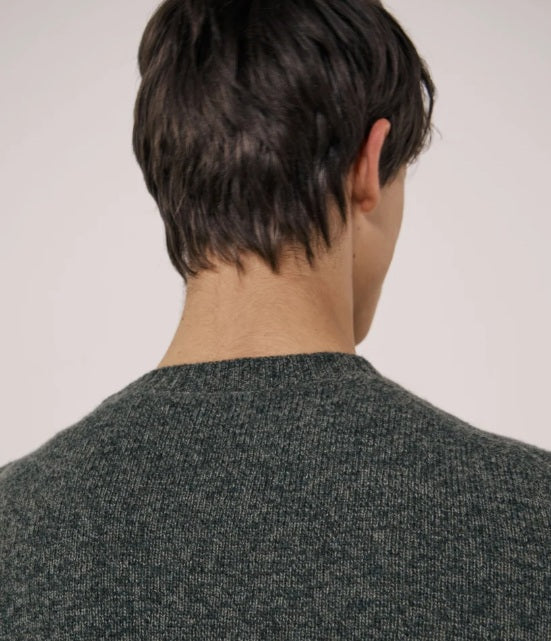 A young man with short dark hair wears the HARTFORD Seamless Wool Crew Pullover in Camo Green and tan pants, standing against a plain light-colored background, looking slightly to the side with a neutral expression.