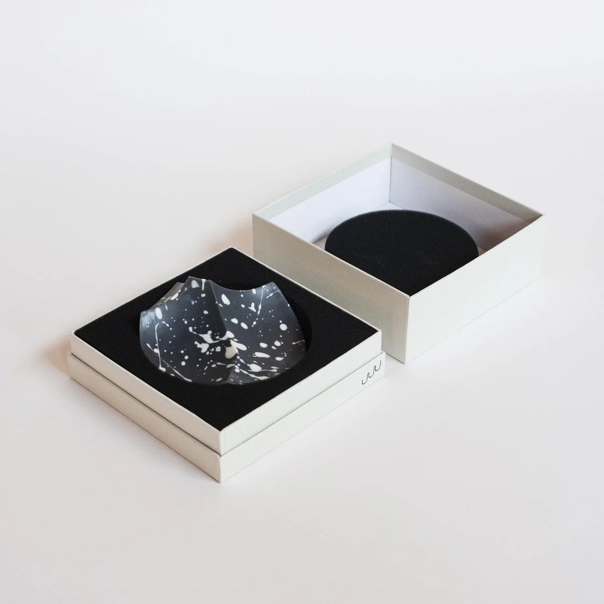 UME STUDIO’s UME - DAILY DETOX ERODE SOAP, a small round black soap with white splatter patterns, is displayed in a square white gift box with black padding. The lid and an extra box with a circular cutout sit beside it on a white surface.