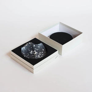 UME STUDIO’s UME - DAILY DETOX ERODE SOAP, a small round black soap with white splatter patterns, is displayed in a square white gift box with black padding. The lid and an extra box with a circular cutout sit beside it on a white surface.