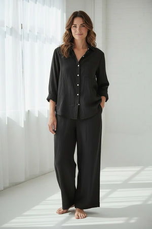A woman with wavy blonde hair stands indoors in a loose black shirt and FRANK & EILEEN’s Maisie Wide Leg Pull On Pant in Washed Linen Black, barefoot on a wooden floor near a chair, vase of green flowers, and staircase.
