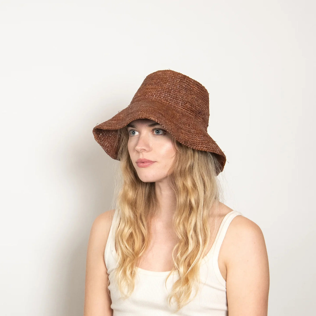 A woman with long, wavy blonde hair wears a white tank top and the WYETH HATS - TALI Raffia Bucket Hat in Chocolate by WYETH, shown in profile against a plain light background.