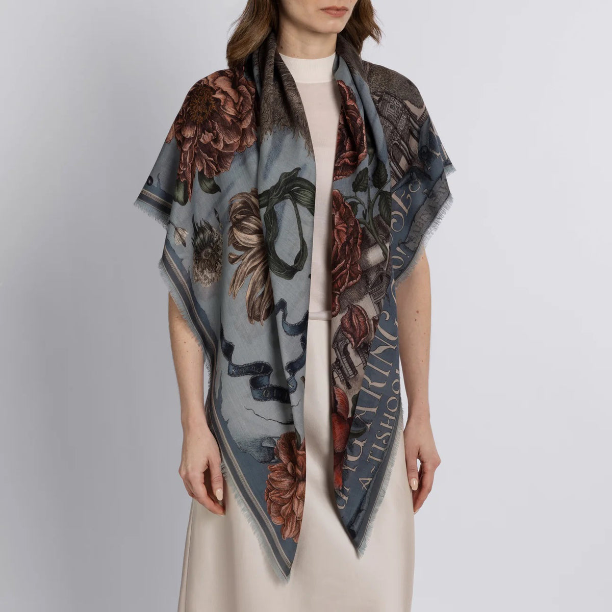A person wearing a light-colored dress drapes the SABINA SAVAGE - RING A RING O' ROSES cashmere scarf in Glacier/Ballet over their shoulders, featuring intricate floral designs reminiscent of English garden flowers. The muted hues contrast beautifully against its soft blue-gray background.