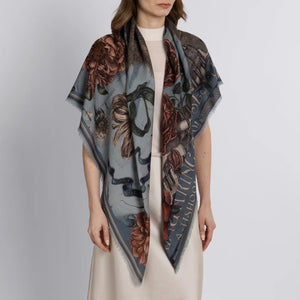 A person wearing a light-colored dress drapes the SABINA SAVAGE - RING A RING O' ROSES cashmere scarf in Glacier/Ballet over their shoulders, featuring intricate floral designs reminiscent of English garden flowers. The muted hues contrast beautifully against its soft blue-gray background.