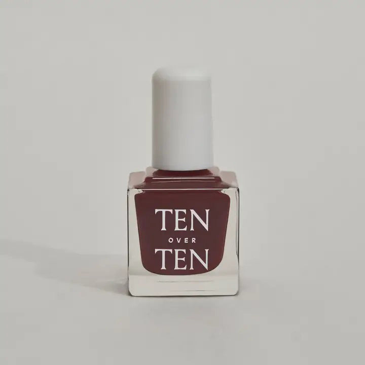A glass bottle of TENOVERTEN - NAIL POLISH from the brand TEN OVER TEN, featuring a white cap and a dusty pink nail color. The front label displays "TEN OVER TEN" in white uppercase letters. This vegan, eight-free formula ensures both beauty and peace of mind. The background is plain and light-colored.