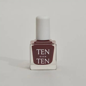 A glass bottle of TENOVERTEN - NAIL POLISH from the brand TEN OVER TEN, featuring a white cap and a dusty pink nail color. The front label displays "TEN OVER TEN" in white uppercase letters. This vegan, eight-free formula ensures both beauty and peace of mind. The background is plain and light-colored.