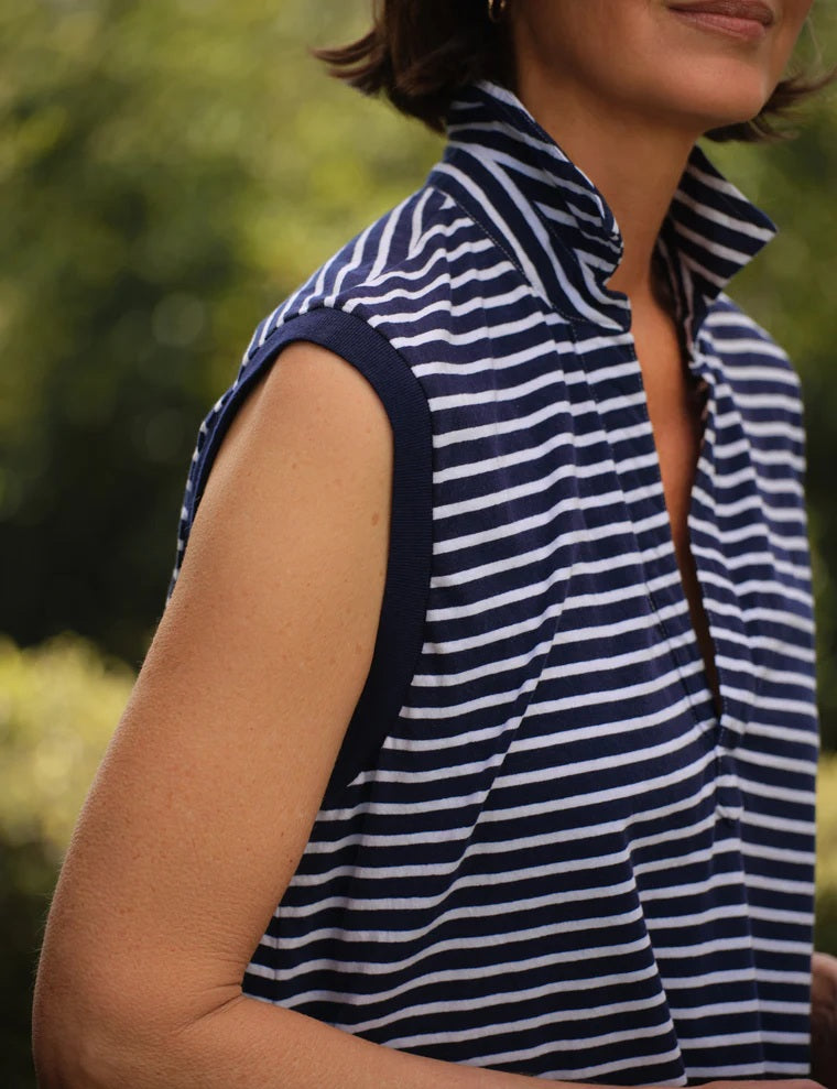 A woman with short brown hair stands outdoors against a backdrop of blurred greenery, wearing the FRANK & EILEEN OLIVIA Popover Henley Tank in Vintage Jersey Yacht Stripe—a sleeveless navy and white striped shirt with a popped collar.