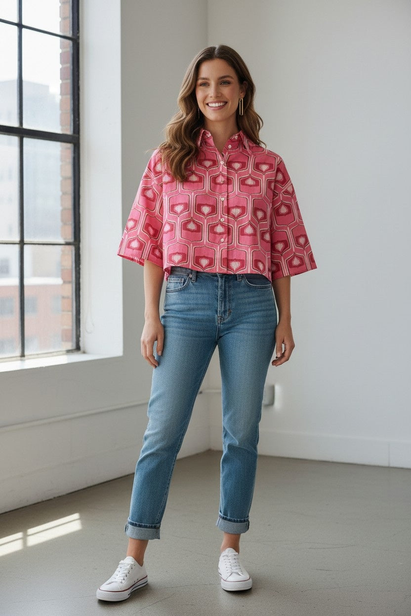 A woman stands indoors by a large window, smiling with long brown hair, and wearing the FLOOR - BOXY CROPPED SHIRT in a pink geometric print from FLOOR, paired with blue cuffed jeans and white sneakers. The stylish cotton top is made in Italy.