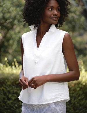 A woman with curly hair stands outdoors in front of green foliage wearing the FRANK & EILEEN Olivia Popover Henley Tank in Famous Denim White, paired with a light-colored striped skirt.