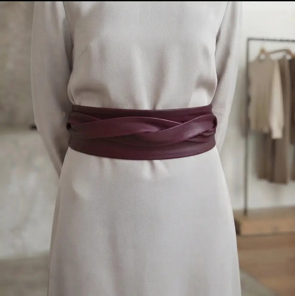An ADA - LEATHER WRAP BELT IN MALBEC, a wide, deep red leather wrap belt worn over a light-colored dress as a versatile accessory.