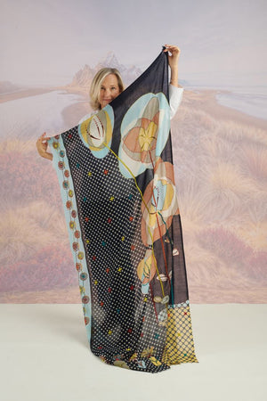 A woman with long blonde hair, in a light blue shirt and dark pants, smiles in front of a scenic backdrop, draping the elegant, hand-embroidered STORIATIPIC - ASTER WOOL SCARF around her shoulders.