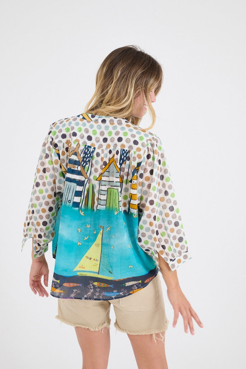 A smiling woman stands with her hands in her pockets, wearing the STORIATIPIC COCO GERTRUDE BLOUSE—featuring polka dot sleeves and a beach hut print—paired with beige shorts, against a plain white background.